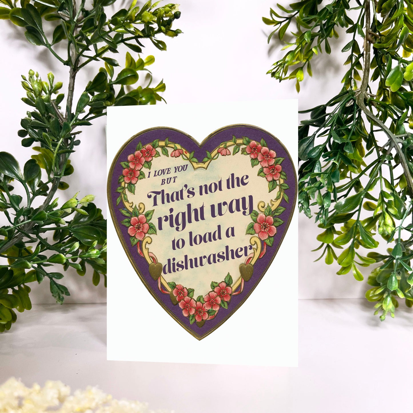 Heart-shaped decorative sign with floral design and thats not how you load a dishwasher text, surrounded by greenery and flowers.