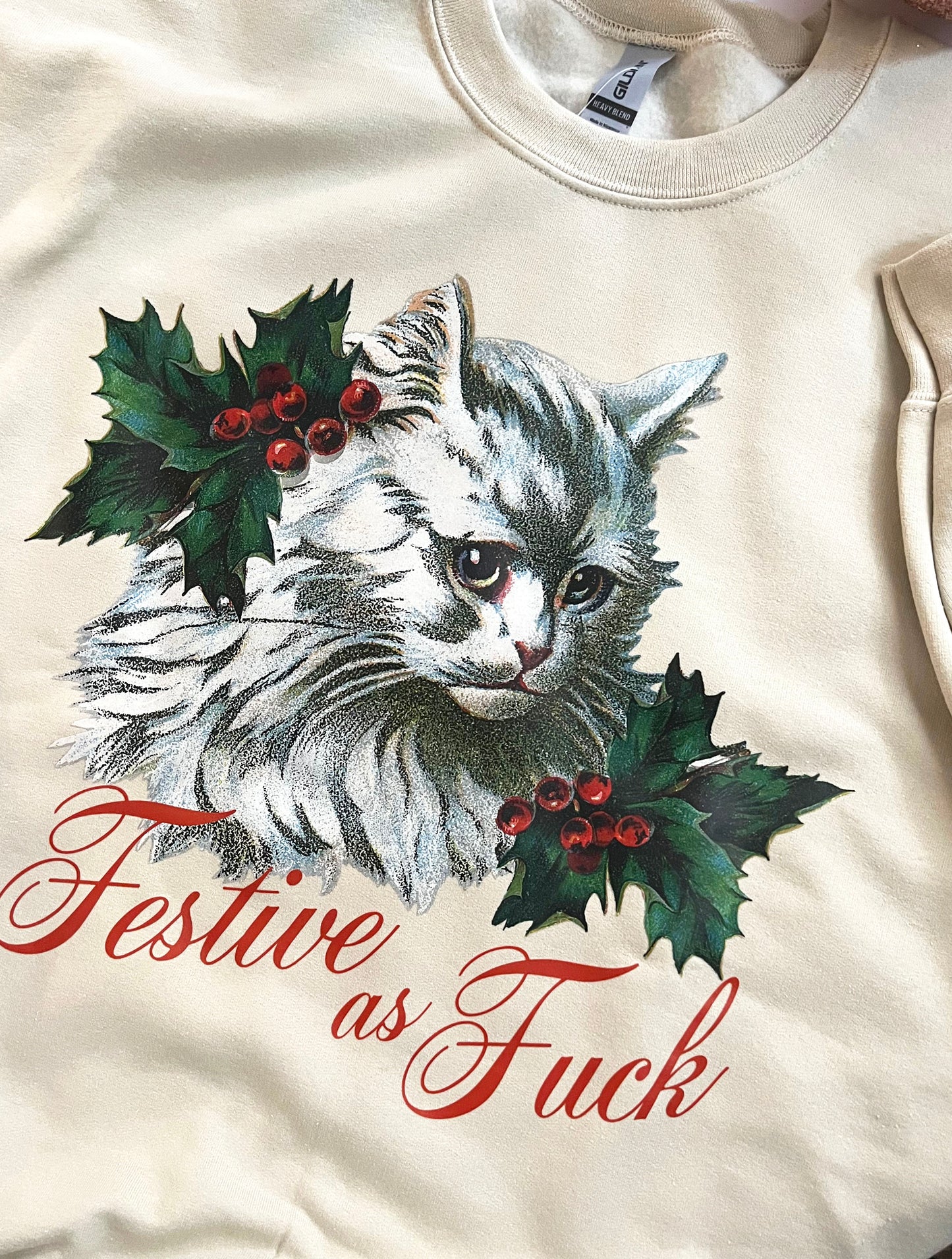 A cream-colored sweatshirt with a graphic of a white cat surrounded by mistletoe with the text 'FESTIVE AS FUCK' below it.