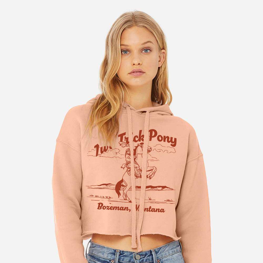 Woman wearing a cropped pink hoodie with cowgirl graphic design and two trick pony text, standing against a plain background