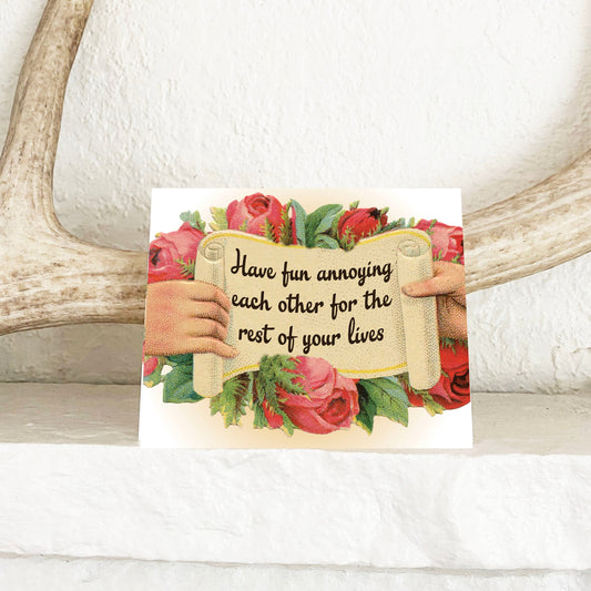 Card with hands holding a scroll with text have fun annoying each other ext and floral design, placed on a wooden surface with antlers.