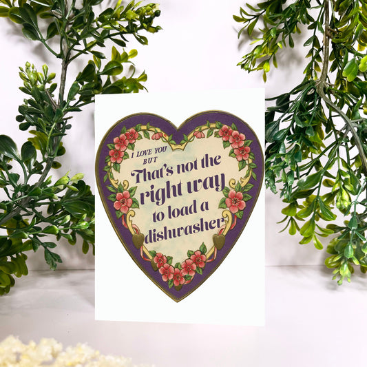 Heart-shaped decorative sign with floral design and thats not how you load a dishwasher text, surrounded by greenery and flowers.