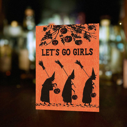 Orange card with black silhouettes of witches and text 'LET'S GO GIRLS' against a blurred bar background.