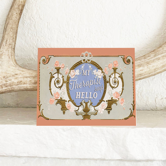 A greeting card with a vintage floral motif and the text 'My Therapist Says Hello' displayed on a white surface, with antique decor in the background.