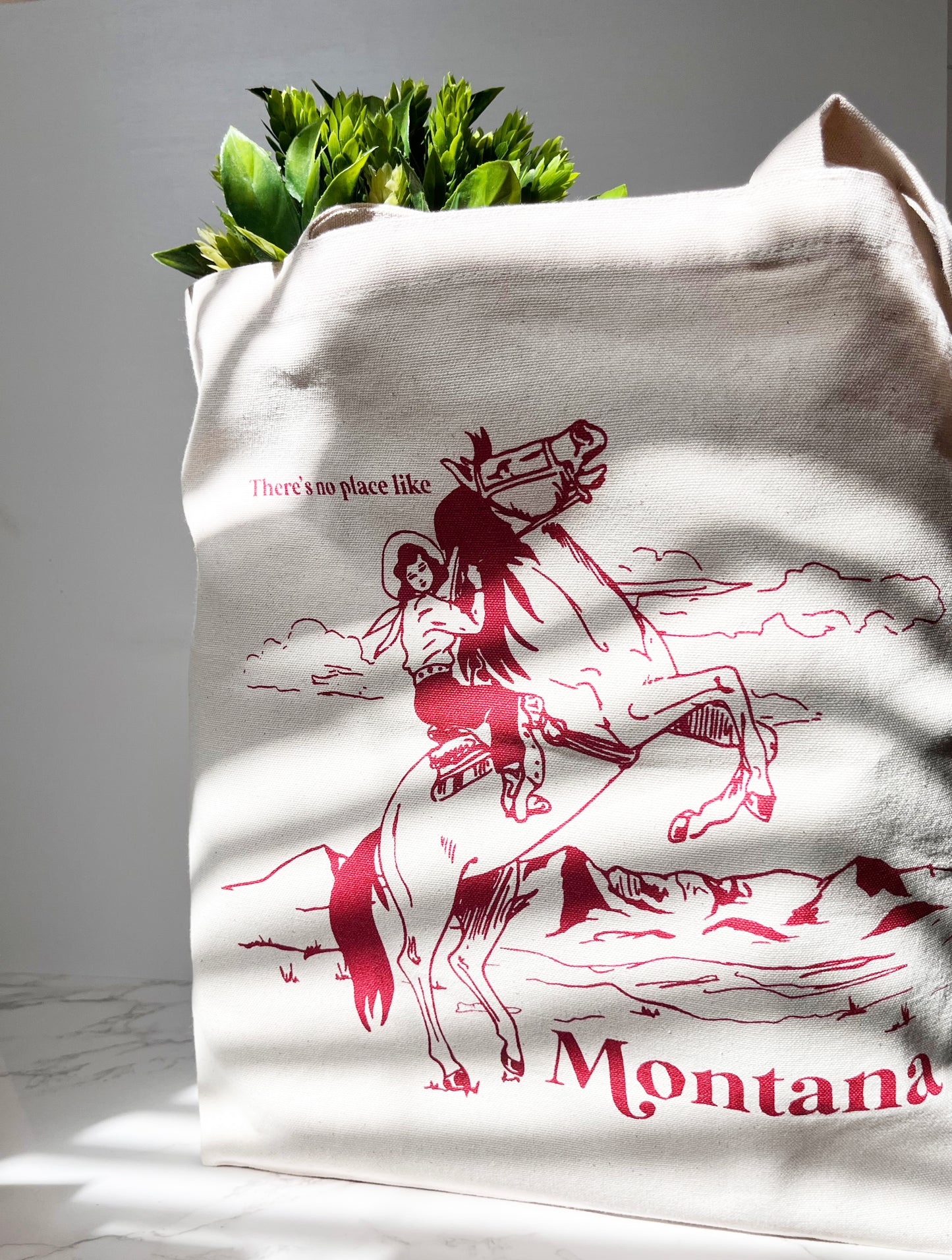 White tote bag with red graphic of a person on a horse and 'Montana' text, filled with green plants.