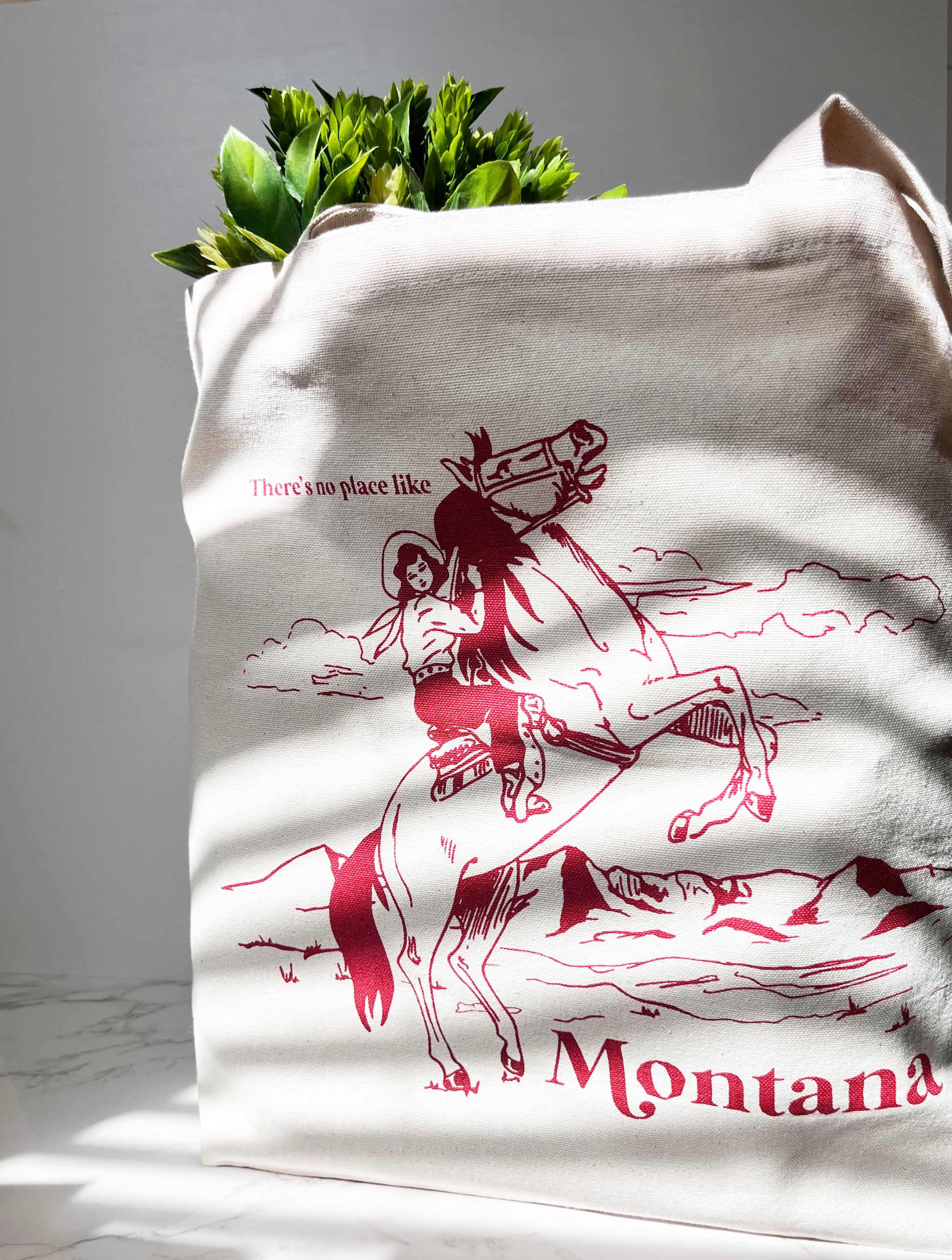 White tote bag with red graphic of a person on a horse and 'Montana' text, filled with green plants.