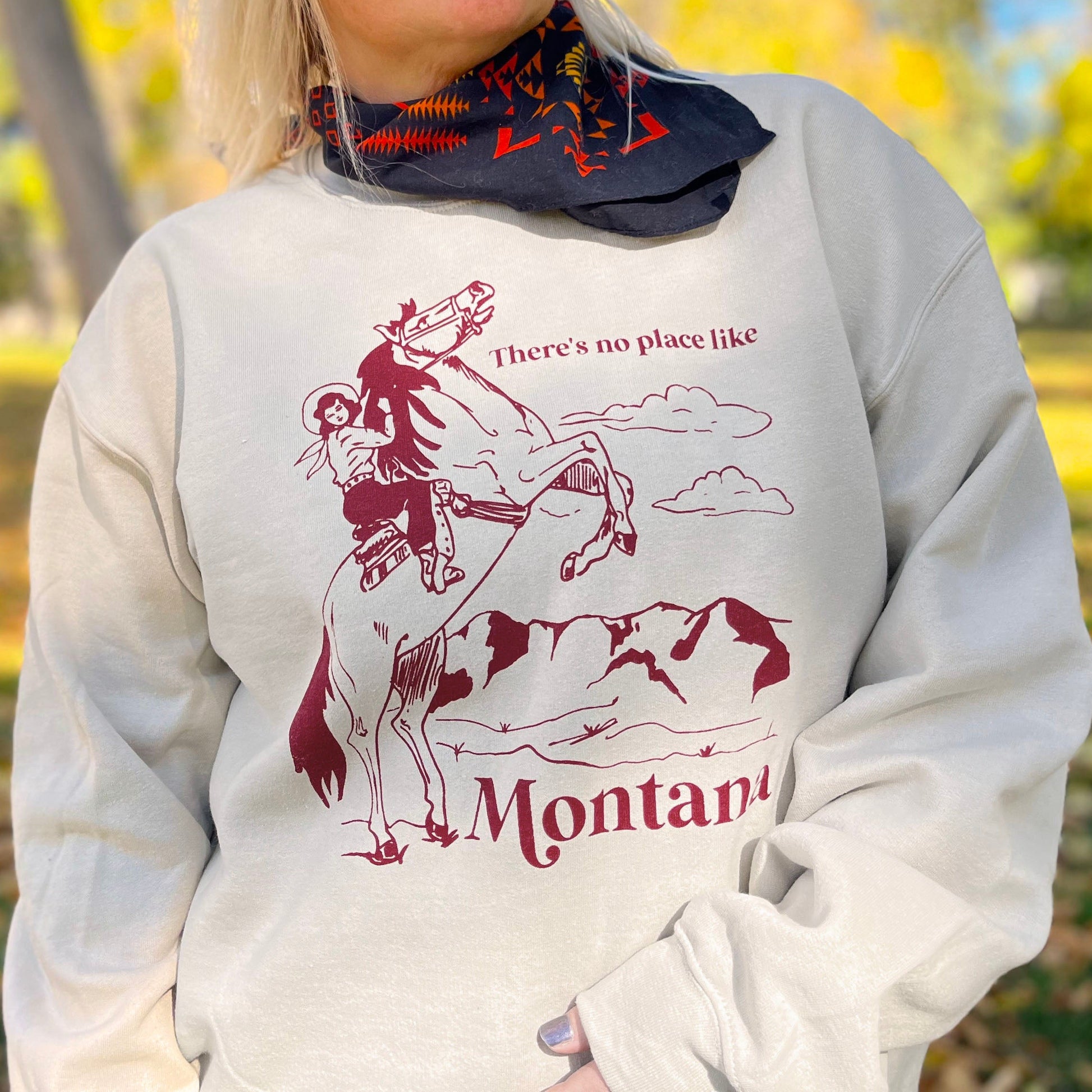 Person wearing a sweatshirt with a graphic design and text that says “There’s no place like Montana” standing outdoors with trees in the background.