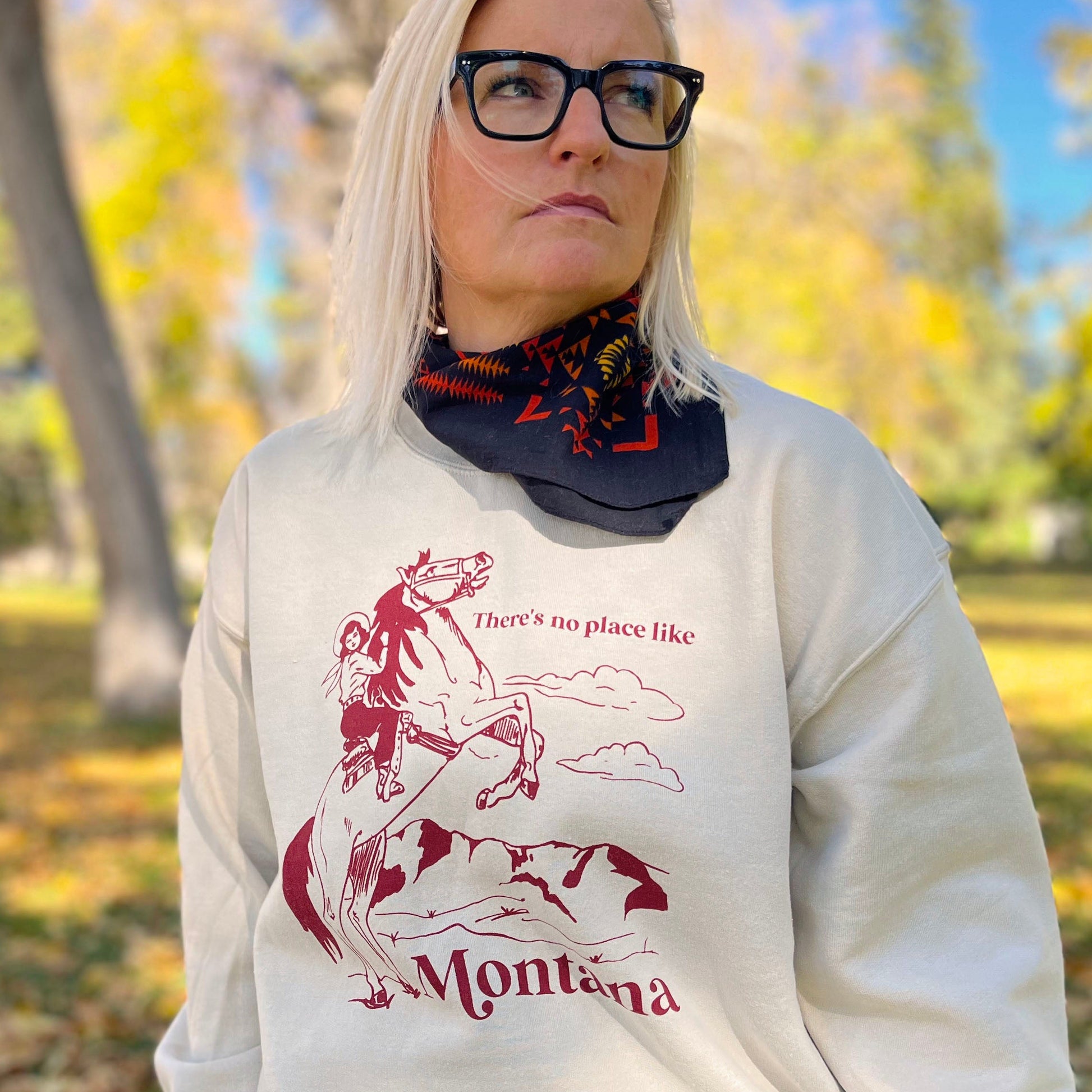 Person wearing a sand colored sweatshirt that says “Theres no place like Montana” text in red with cowgirl on a horse on it.