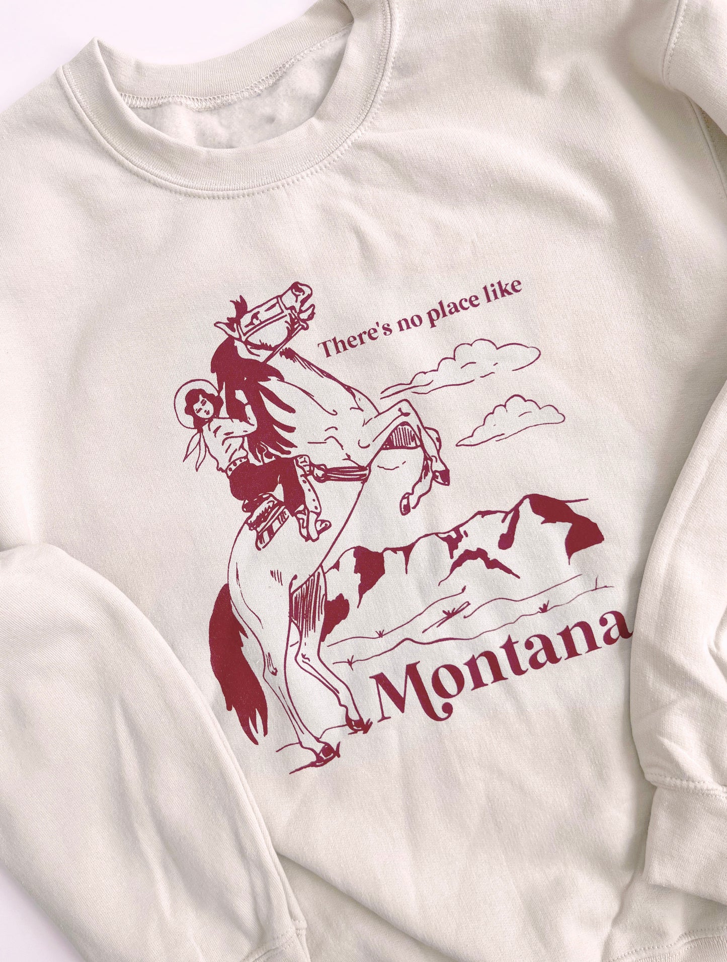 Sand colored sweatshirt that says “There’s no place like Montana” in red text with a cowgirl on a horse on it. 