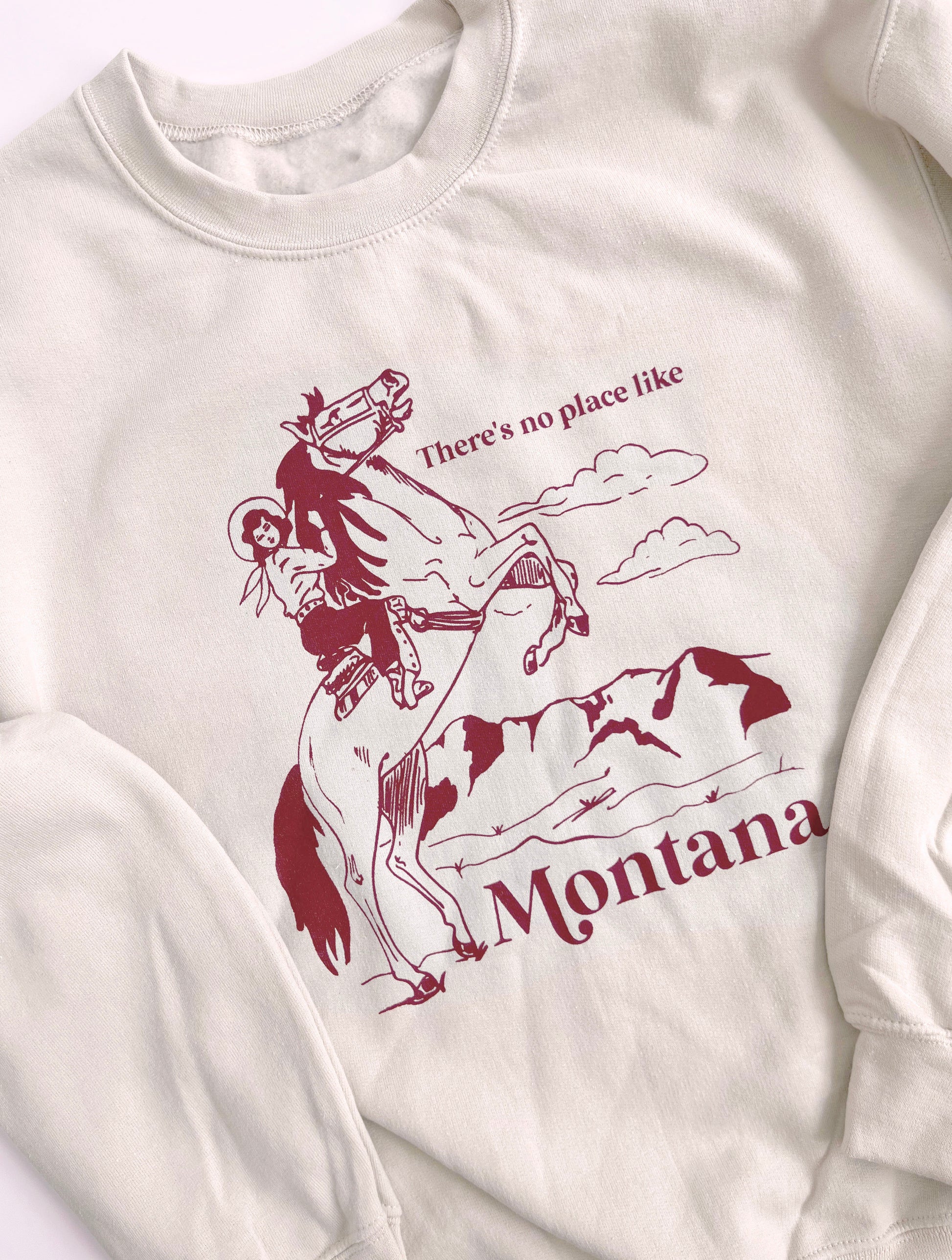 Sand colored sweatshirt that says “There’s no place like Montana” in red text with a cowgirl on a horse on it. 