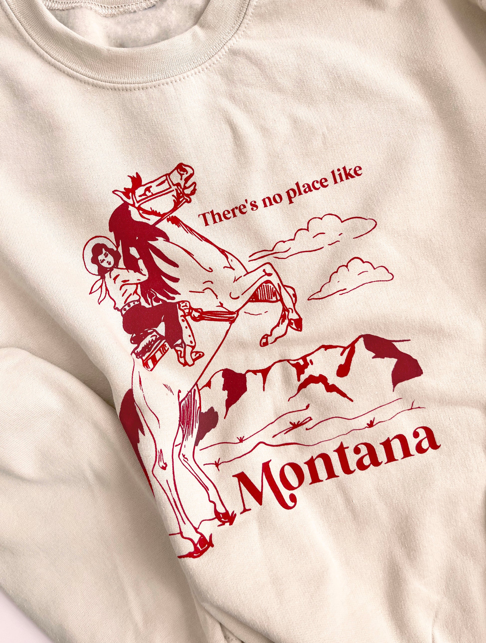 Sand colored sweatshirt that says “There’s no place like Montana” in red text with a cowgirl on a horse on it. 