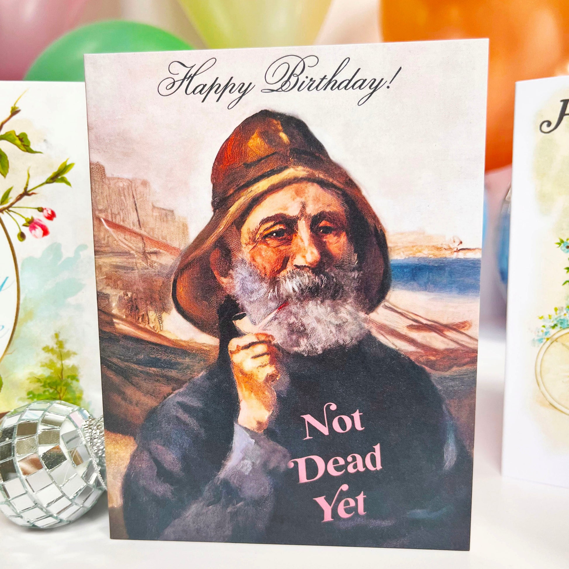 Birthday card with a humorous message 'Not Dead Yet' featuring a person in a hat, surrounded by colorful balloons.