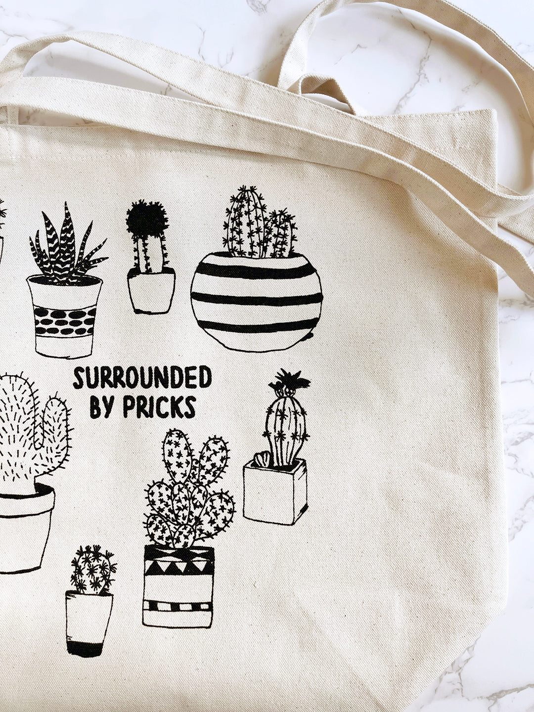 Beige tote bag with black illustrations of potted plants and text 'Surrounded by Pricks' on a marble surface.